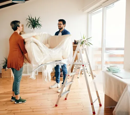 Two people carrying a sofa wrapped in plastic into a flat with large windows